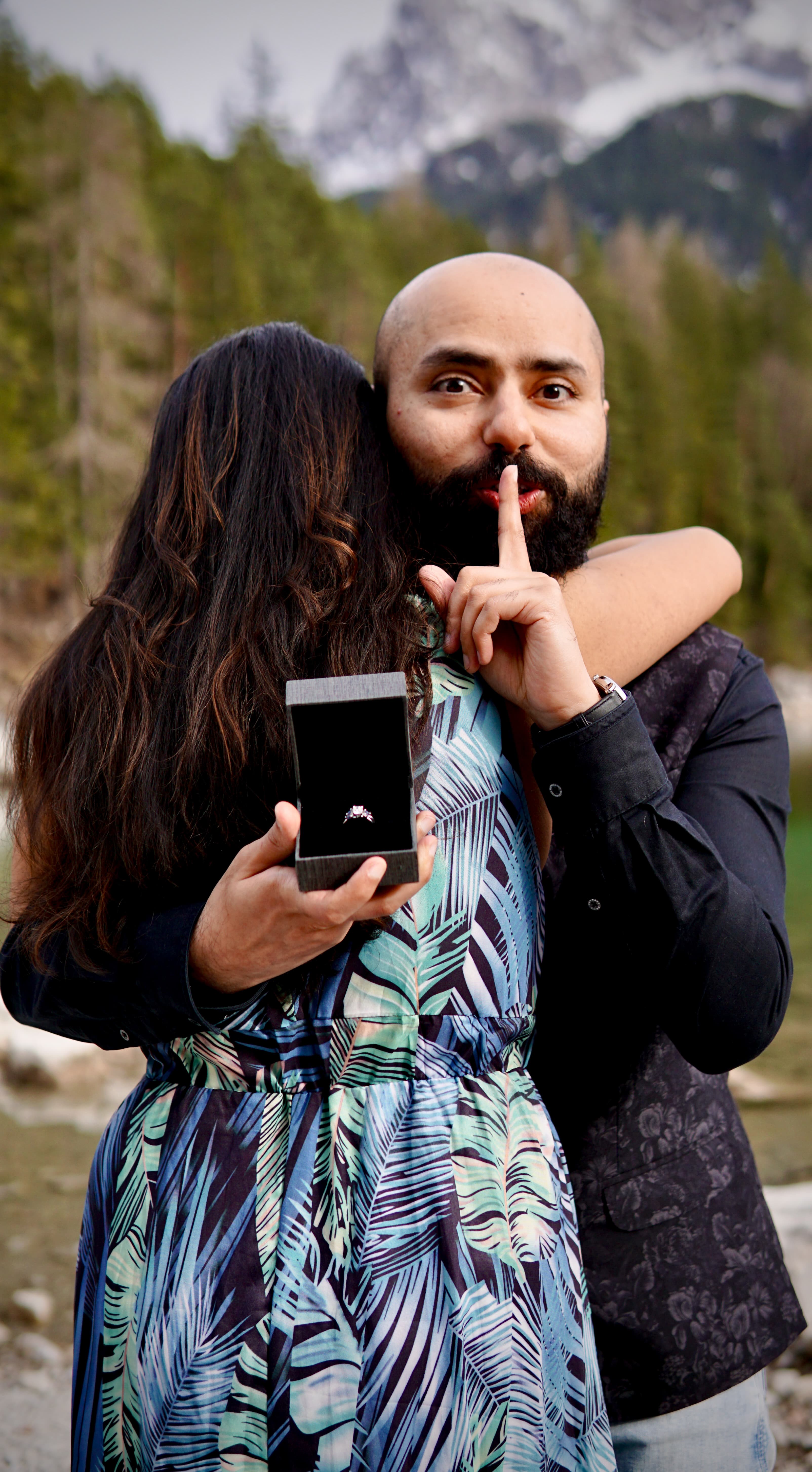 Man holding a ring box behind his partner with a finger on his lips in a surprise proposal moment, pine forest and snowy peaks behind